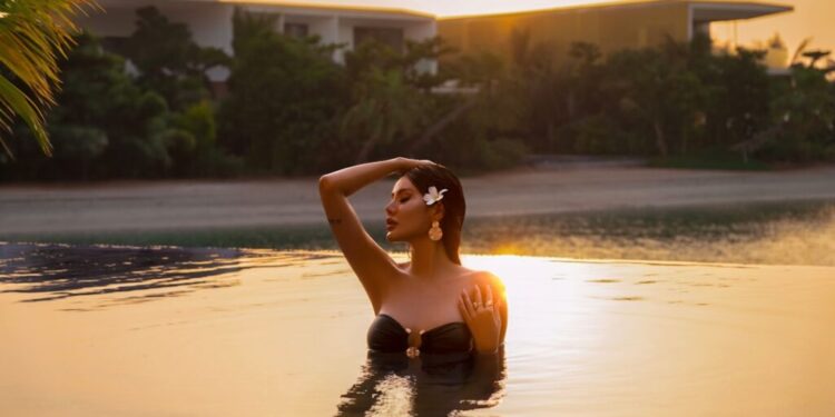 Woman in a black bikini standing chest-deep in a sunset-lit pool, hand resting on her head with a flower in her hair.