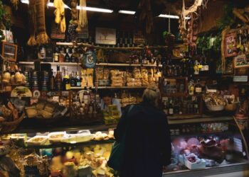 Cozy market stall with shelves of bottles, jars, and dried goods; a customer browses the counter display behind glass.