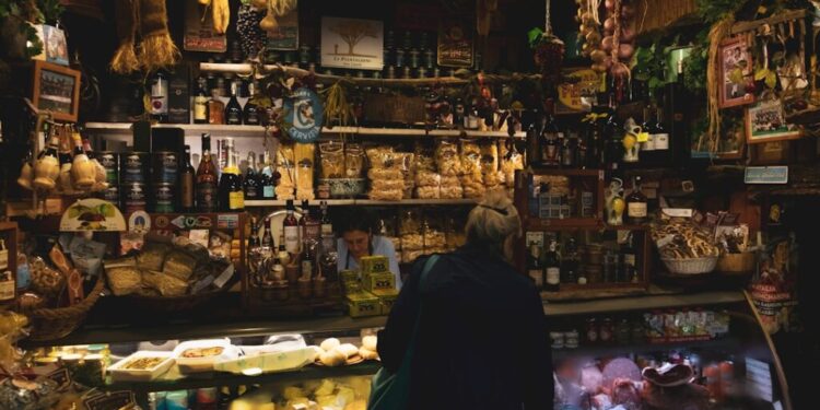 Bancarella accogliente del mercato con scaffali di bottiglie, barattoli e prodotti secchi; un cliente sfoglia il display del bancone dietro il vetro.