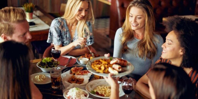 レストランで一緒にダイニングしている友人のグループ。彼らは大きなお皿の食べ物をテーブルの横で共有して渡しながら笑っています。