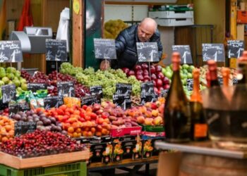 Colorful fruit stall at a market with a worker behind the display and chalkboard price signs visible among the produce.