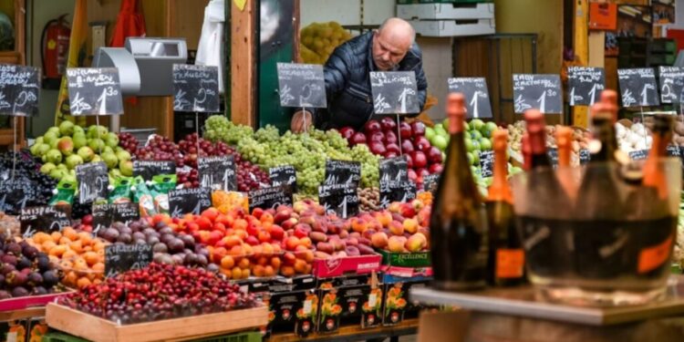 Farbenfroher Obst- und Gemüsestand auf einem Markt mit einem Arbeiter hinter der Auslage und Tafeln mit Preisen zwischen den Produkten.
