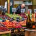 Colorful fruit stall at a market with a worker behind the display and chalkboard price signs visible among the produce.