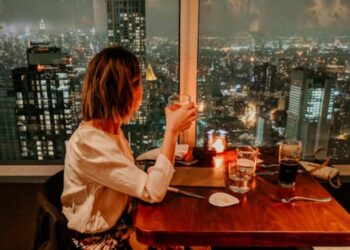 Woman with short hair in a white blouse sits at a wooden table by a window, holding a drink, overlooking a city skyline at night with lit buildings and candles on the table.