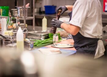Chef in a white uniform and gloves grinds pepper onto meat at a busy kitchen prep station, with greens and utensils around.