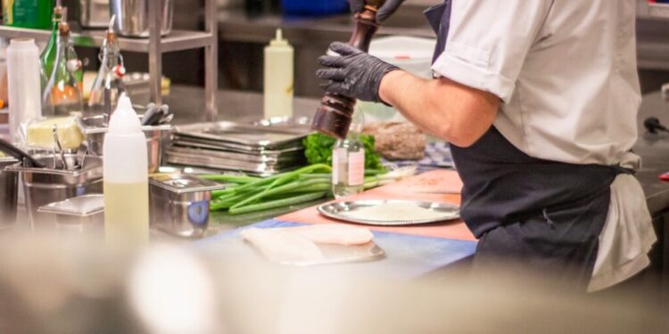 Chef in a white uniform and gloves grinds pepper onto meat at a busy kitchen prep station, with greens and utensils around.