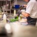 Chef in a white uniform and gloves grinds pepper onto meat at a busy kitchen prep station, with greens and utensils around.