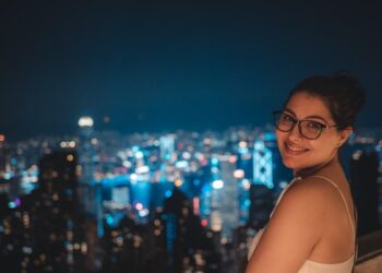 Smiling woman with glasses on a rooftop at night, city skyline blurred in the background.