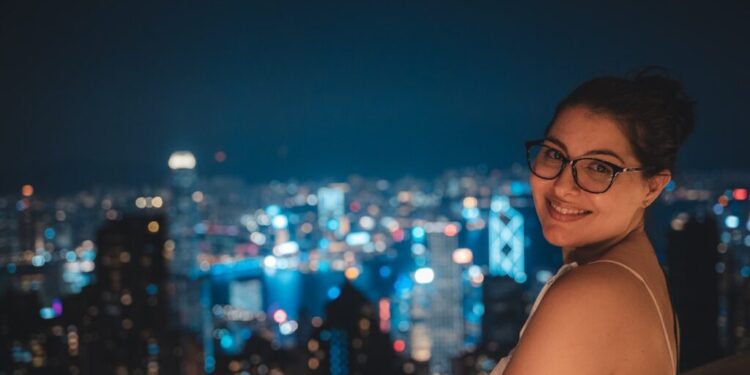 Dachbars in Tokio – jeder Yen wert - GUSTOwebzine Lächelnde Frau mit Brille auf einem Dach in der Nacht, Skyline der Stadt verschwommen im Hintergrund.