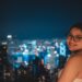 Lächelnde Frau mit Brille auf einem Dach in der Nacht, Skyline der Stadt verschwommen im Hintergrund.