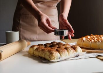 Baker presses a round metal cookie cutter into dough balls on a white countertop, with a rolling pin and two trays of bread buns nearby.
