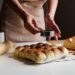 Baker presses a round metal cookie cutter into dough balls on a white countertop, with a rolling pin and two trays of bread buns nearby.