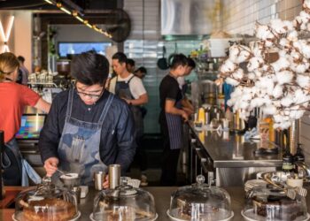 Barista in a denim apron scooping dessert at a busy cafe counter with glass cake domes in the foreground.