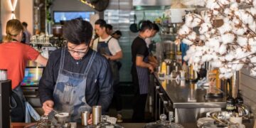 Barista in a denim apron scooping dessert at a busy cafe counter with glass cake domes in the foreground.