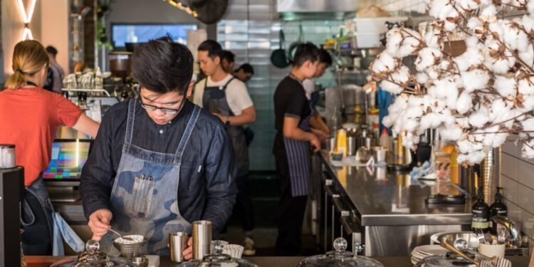 Barista in a denim apron scooping dessert at a busy cafe counter with glass cake domes in the foreground.