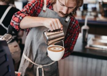 Barista in a plaid shirt pours steamed milk from a metal pitcher into a latte in a white cup at a cafe