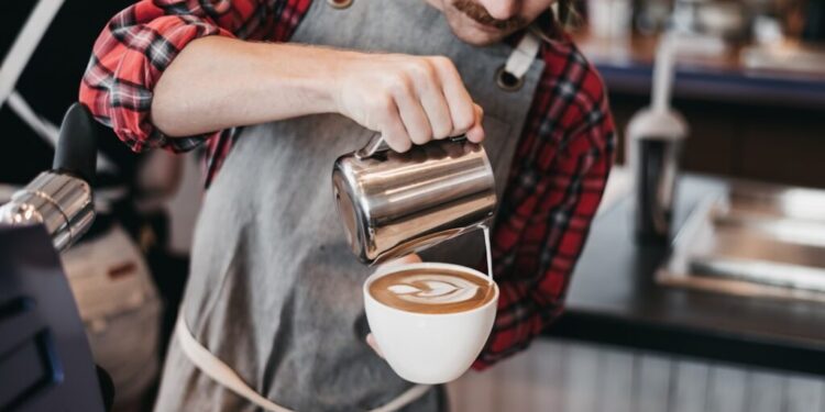 Barista in a plaid shirt pours steamed milk from a metal pitcher into a latte in a white cup at a cafe