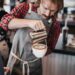 Singapore coffee championship what to expect - GUSTOwebzine Barista in a plaid shirt pours steamed milk from a metal pitcher into a latte in a white cup at a cafe