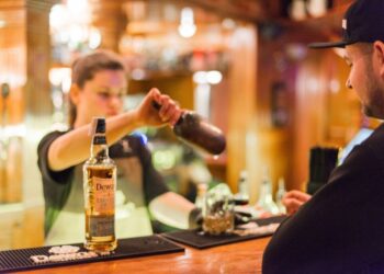 Bartender pouring whiskey from a bottle into a shaker for a customer at a bar by the counter.