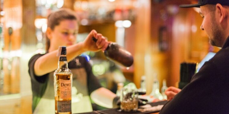 Bartender pouring whiskey from a bottle into a shaker for a customer at a bar by the counter.
