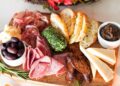 a colorful summer grazing board with global pantry staples including cured meats, cheese, and fresh fruit on a wooden surface