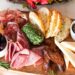 a colorful summer grazing board with global pantry staples including cured meats, cheese, and fresh fruit on a wooden surface