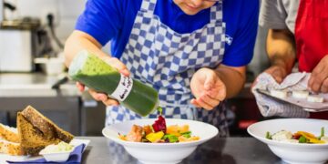 A chef carefully pouring chilled soups into a white bowl in a professional kitchen