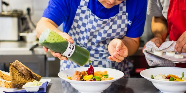 A chef carefully pouring chilled soups into a white bowl in a professional kitchen