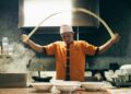 Chef in orange uniform tossing dough in a busy kitchen with steam and bowls on the counter behind him