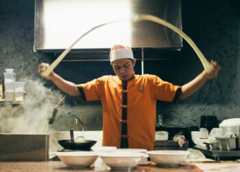 Chef in orange uniform tossing dough in a busy kitchen with steam and bowls on the counter behind him