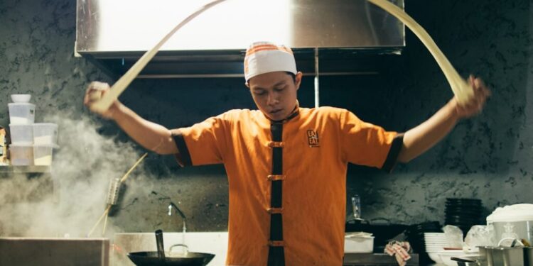 Chef in orange uniform tossing dough in a busy kitchen with steam and bowls on the counter behind him