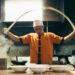 Chef in orange uniform tossing dough in a busy kitchen with steam and bowls on the counter behind him