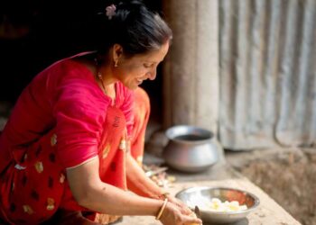 Woman in a bright pink saree chops vegetables in a rustic outdoor kitchen, with metal bowls nearby.