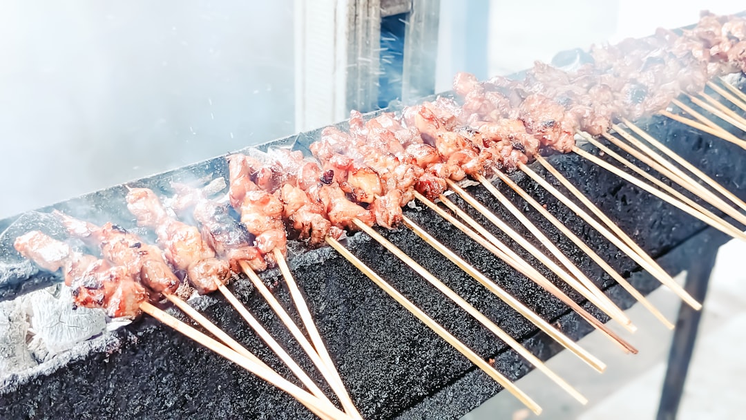A vendor grilling street food satay skewers over charcoal at an outdoor market stall, with smoke rising around the meat.