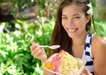 Smiling woman outdoors with a white flower in her hair holding a bowl of colorful shaved ice dessert on a spoon.