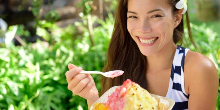 Kakigori a Tokyo 7 negozi specializzati - GUSTOwebzine Donna sorridente all'aperto con un fiore bianco tra i capelli che tiene una ciotola di dessert di ghiaccio tritato colorato su un cucchiaio.