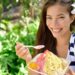 Smiling woman outdoors with a white flower in her hair holding a bowl of colorful shaved ice dessert on a spoon.