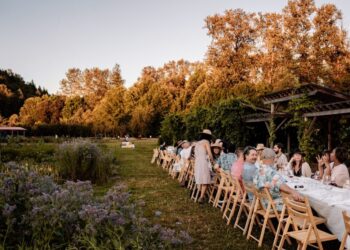 Long outdoor garden party or wedding reception with a long table of guests dining under a wooden pergola as autumn trees glow in the background