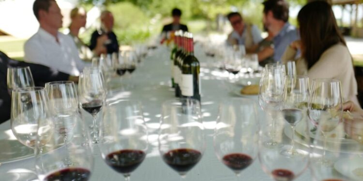 EU wijngaardervaring om nu te boeken - GUSTOwebzine Al fresco-lunch buiten met een lange tafel, wijnglazen en flessen terwijl gasten onder bomen spreken.