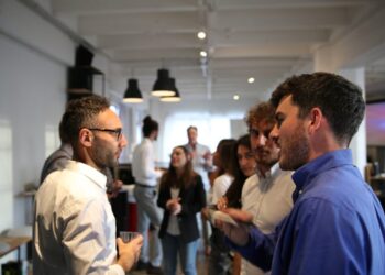 Group of coworkers networking at an office party, some holding drinks and chatting in a bright open space with others in the background.]