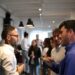 Gruppo di colleghi che si conoscono durante un evento in ufficio, alcuni con drink in mano e che chiacchierano in uno spazio aperto e luminoso con altri sullo sfondo.