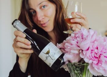 Woman holding a wine bottle and glass, smiling, with pink flowers in a vase nearby as she pours wine.
