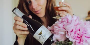 Woman holding a wine bottle and glass, smiling, with pink flowers in a vase nearby as she pours wine.