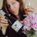 Woman holding a wine bottle and glass, smiling, with pink flowers in a vase nearby as she pours wine.