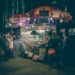 Nighttime street food cart with illuminated signs and steam rising as customers queue nearby on a city sidewalk.