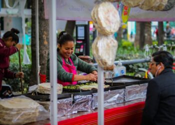 Street food vendor flipping food on griddles at a busy outdoor cart, with customers seated nearby under colorful awnings.