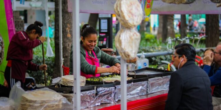 Street food vendor flipping food on griddles at a busy outdoor cart, with customers seated nearby under colorful awnings.