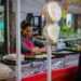 Street food vendor flipping food on griddles at a busy outdoor cart, with customers seated nearby under colorful awnings.