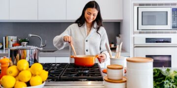 Smiling woman stirs an orange pot on a gas stove in a bright, modern kitchen with lemons on the counter in front of her