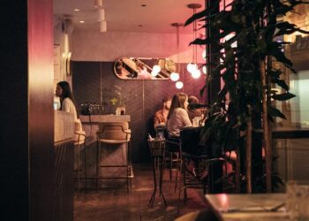 Café interior at night with warm pink lighting, patrons seated at tables near a bar; leafy plants in the foreground.
